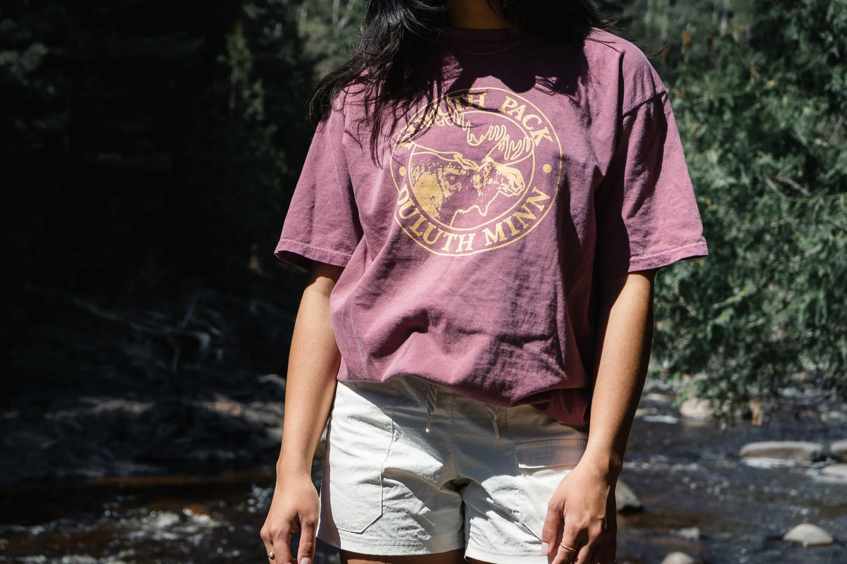 Maroon Duluth Pack logo t shirt being worn by a woman in white shorts. There is a river and pine trees in the background