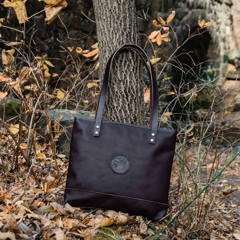 Leather Lakewalk Tote in dark premium leather with riveted handles, resting against a tree in a rustic autumn forest setting.