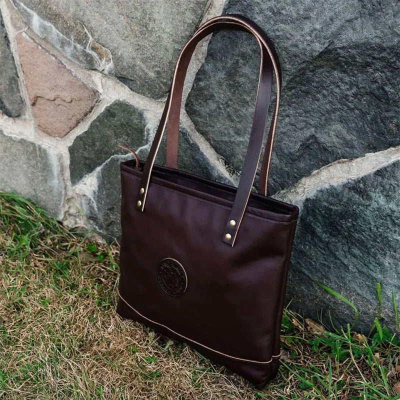Leather Lakewalk Tote in rich brown leather with riveted handles, set outdoors against a stone wall, highlighting durable craftsmanship and rugged style.