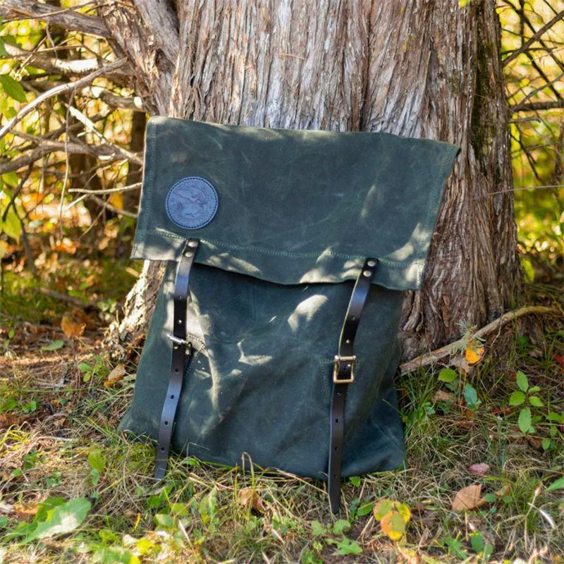 #51 Utility Pack in dark green waxed canvas with premium leather straps, resting against a tree outdoors, showcasing durable craftsmanship for rugged use.
