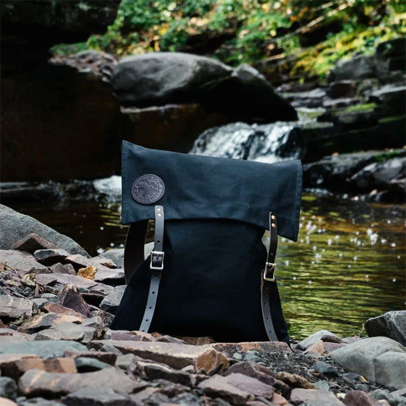 #51 Utility Pack in black tough 15-ounce canvas with premium leather straps, set on rocky terrain by a flowing mountain stream, highlighting durable outdoor use.