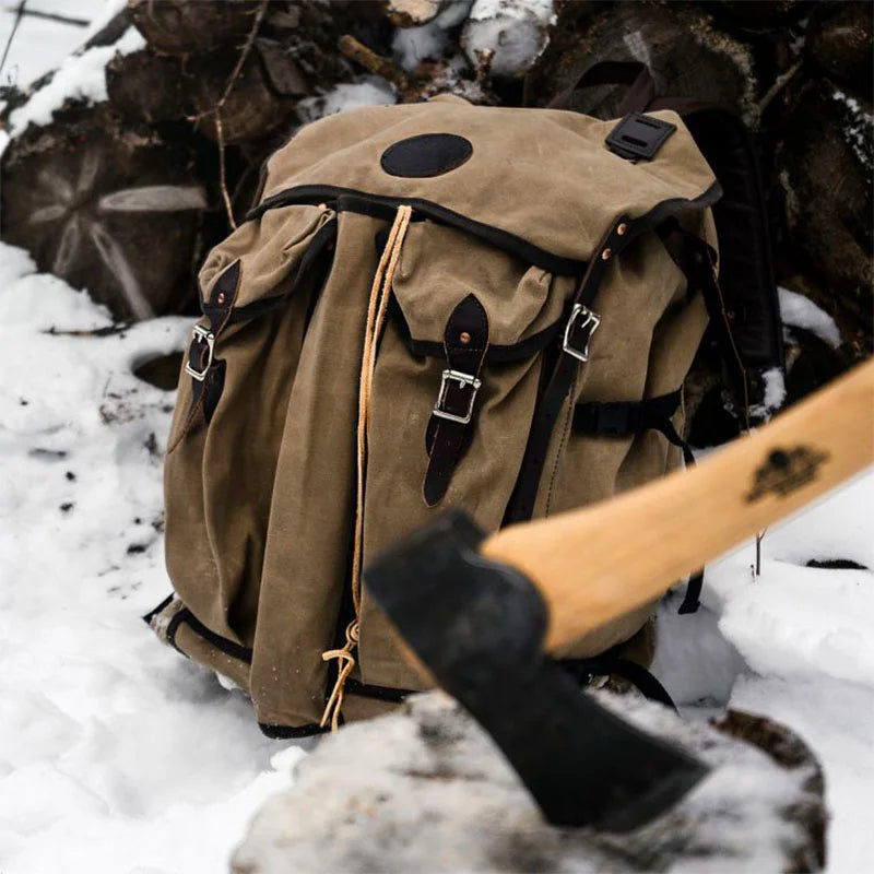 Bushcrafter backpack in rugged canvas with leather straps resting on snow near chopped firewood and an axe, highlighting durable outdoor gear by Duluth Pack.