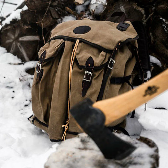 Bushcrafter backpack in rugged canvas with leather straps resting on snow near chopped firewood and an axe, highlighting durable outdoor gear by Duluth Pack.