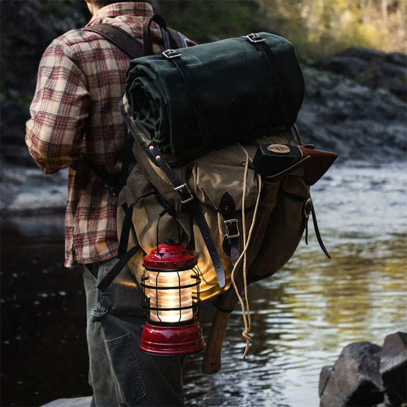 Bushcrafter rugged 15-ounce canvas backpack with leather accents, featuring rolled green canvas bedroll and hanging lantern, worn by outdoorsman by riverbank