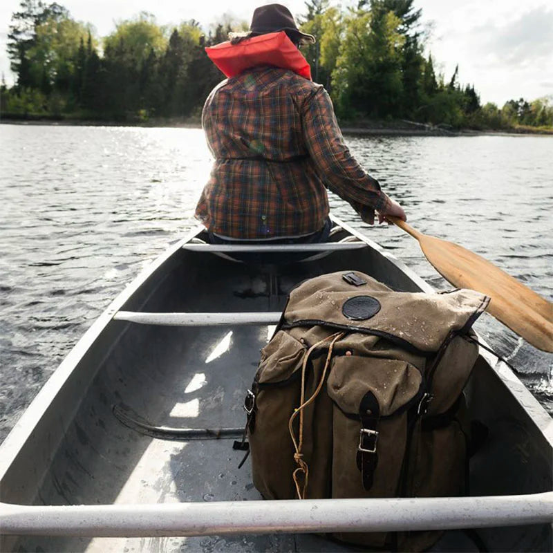 Bushcrafter backpack in rugged 15-ounce canvas sits in a canoe while a person paddles on a calm lake surrounded by trees, illustrating durable outdoor gear.