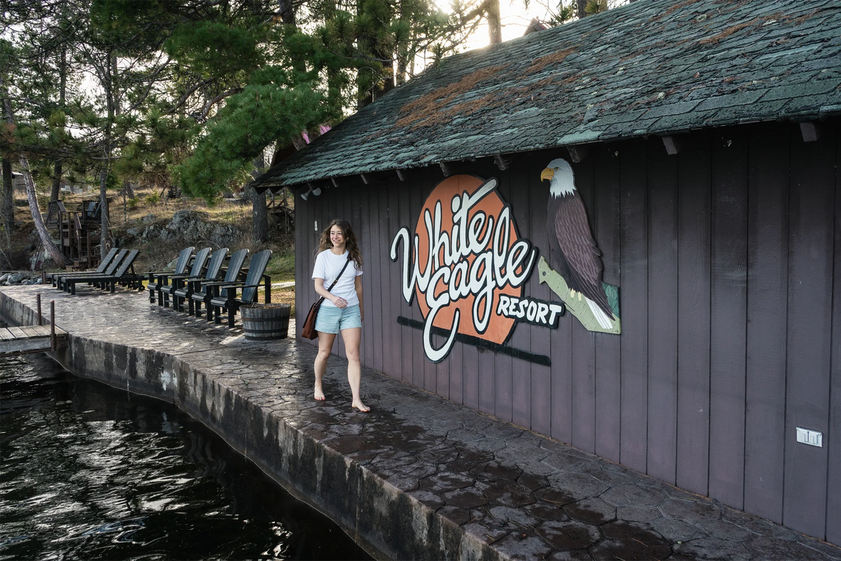 Woman walking on a dock in the summer in front of a White Eagle Resort sign. She is wearing a Duluth Pack Bison Leather Haversack. 