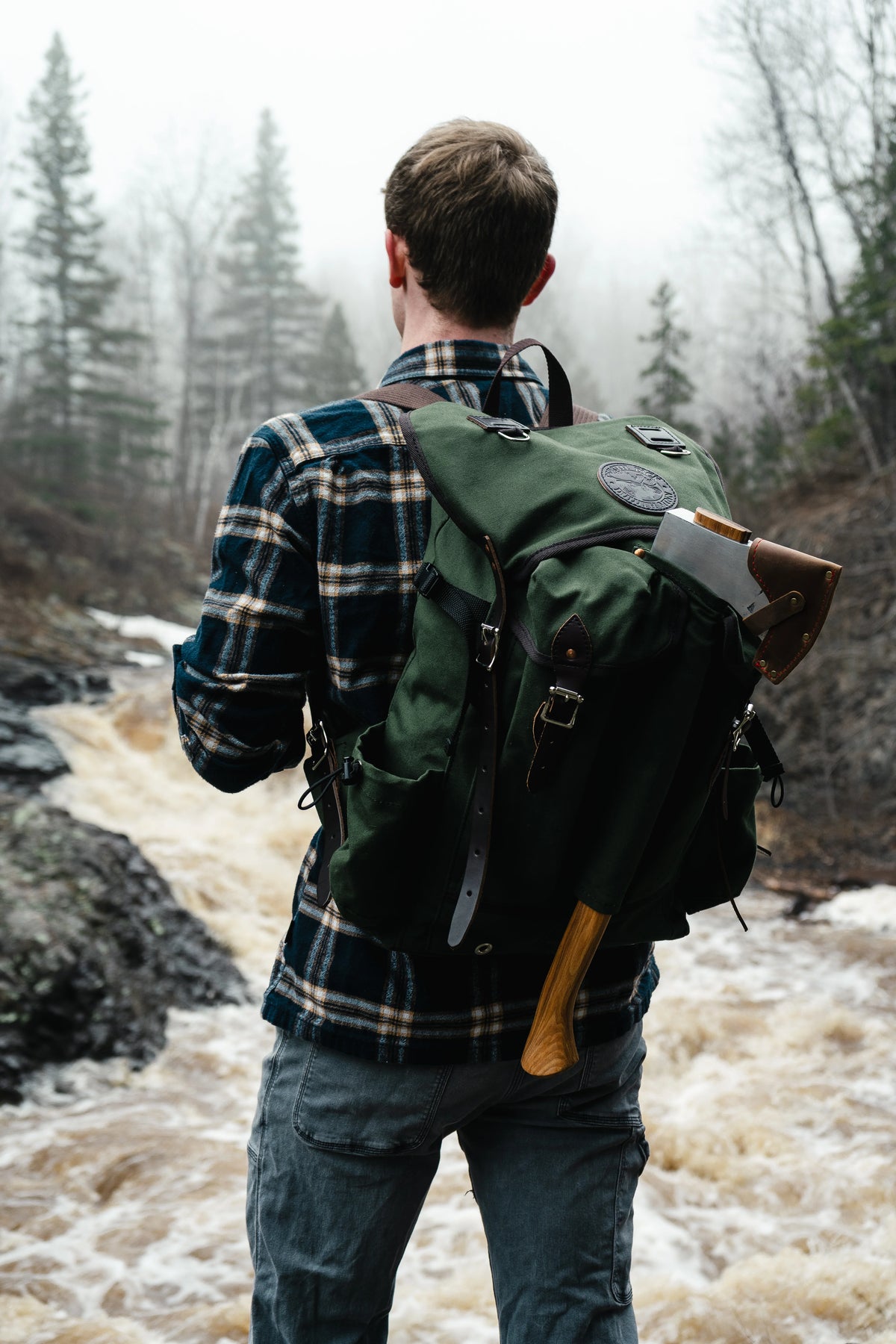 Duluth Pack X Brant and Cochran bundle showing a man wearing a olive drab Outlander Pack standing in front of a waterfall. 