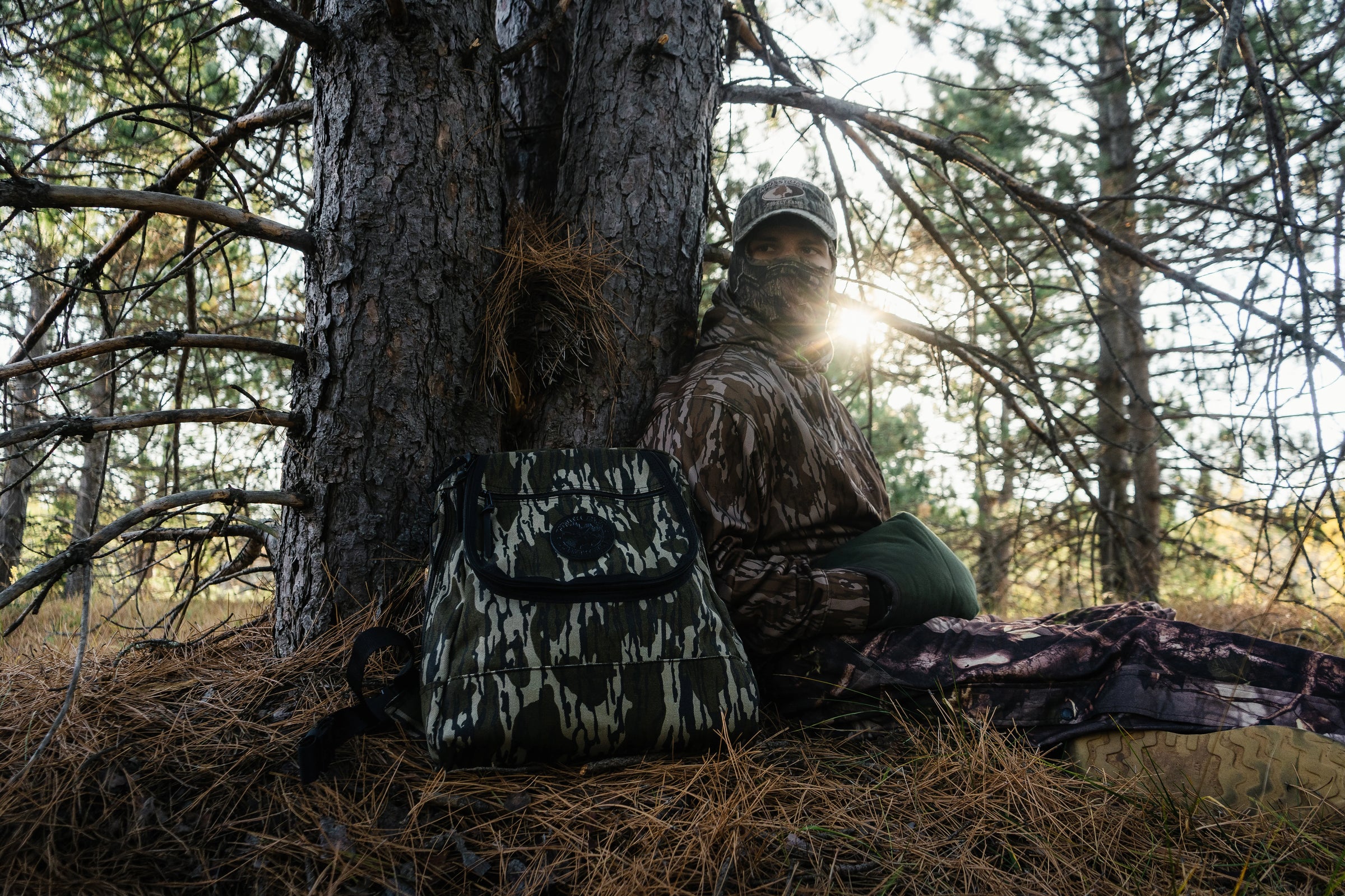 Handmade canvas hunting backpack with camouflage pattern placed against a tree in a forest setting beside a person dressed in full camouflage gear, highlighting outdoor durability and heritage craftsmanship.