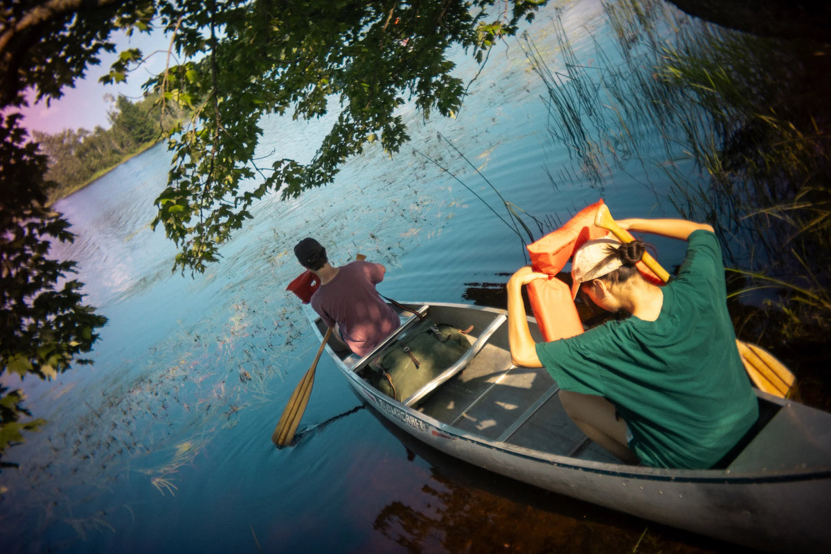 A man and woman in a aluminum canoe putting on life jackets to paddle in the BWCA. They have a #2 Original Duluth Pack Canoe pack. 
