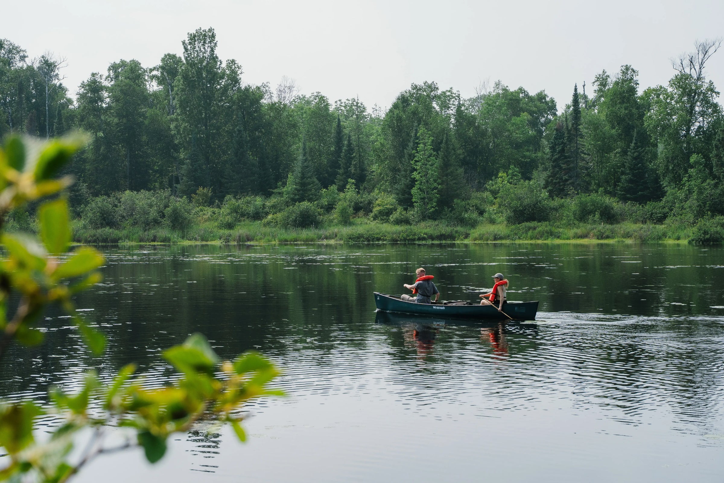 Photo: A man and woman canoeing on a clear lake. They are surrounded by a green forest. 