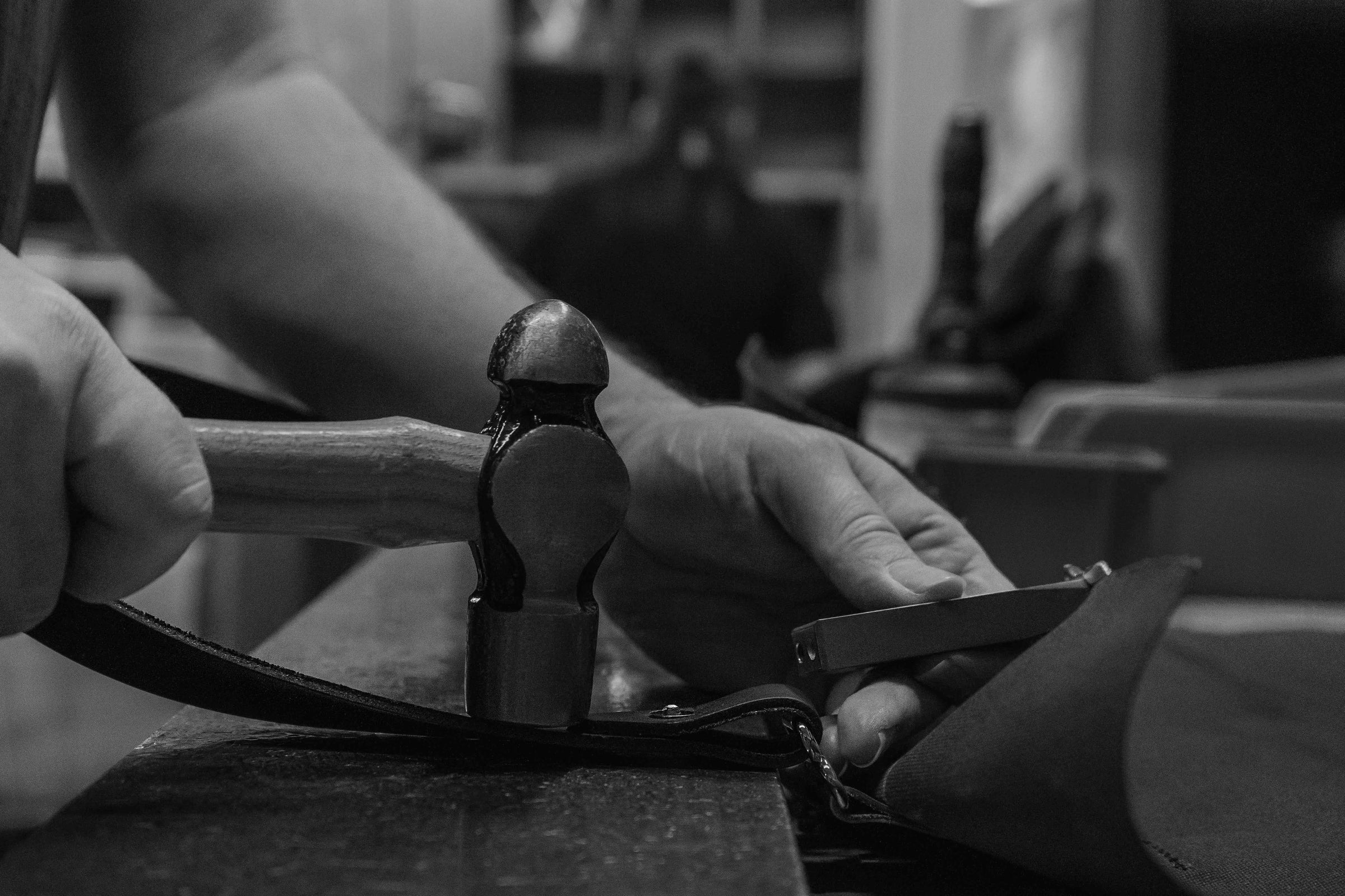 Man hammering a rivet into a piece of leather on a handmade leather good with a workshop in the background
