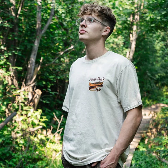 Duluth Pack Canoe Adventure T-Shirt in cappuccino, featuring outdoor canoe graphics, worn by a man in a forest setting, made by LAKESHIRTS, INC.
