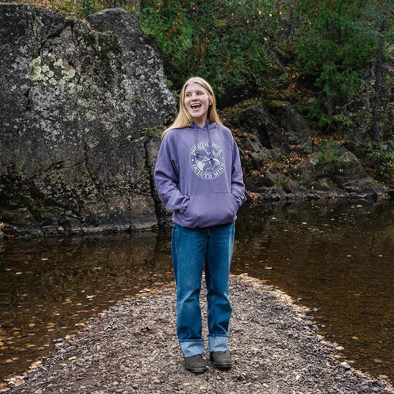 A young woman standing on a rocky path surrounded by shallow water, wearing a purple Duluth Pack Logo Sweatshirt, blue cuffed jeans, and dark shoes, smiling and looking off to the side with a natural forest and rock background.