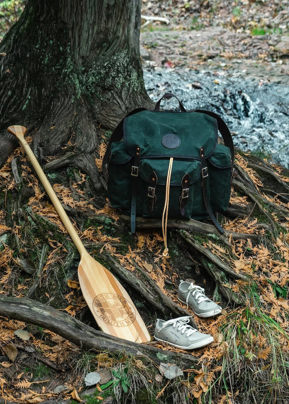 An image of a Duluth Pack Gitchi Gami Wanderer hiking backpack with a Bending Branches BB Special 11 Canoe Paddle and a pair of Loyak barefoot Astral shoes. The products are cedar tree roots with a waterfall in the background.