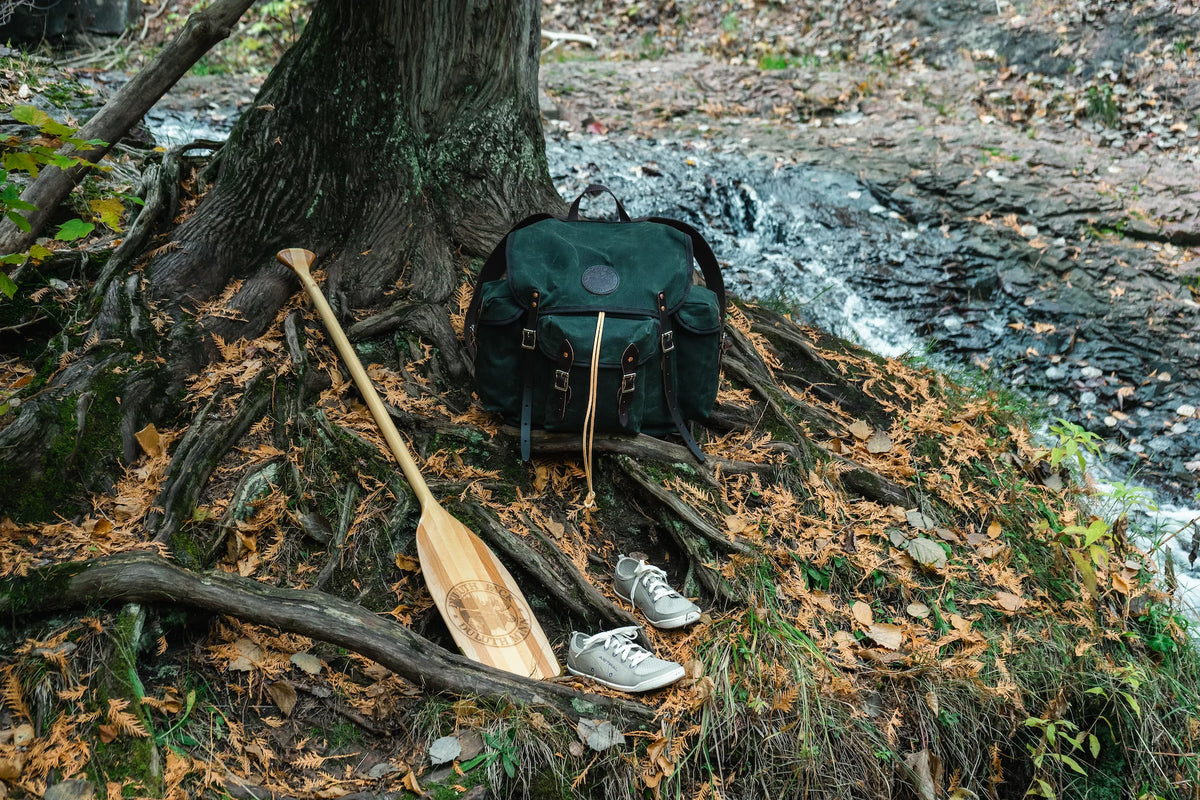 An image of a products won in the Outdoorsman's Giveaway. Win a Duluth Pack Gitchi Gami Wanderer hiking backpack with a Bending Branches BB Special 11 Canoe Paddle and a pair of Loyak barefoot Astral shoes. The products are cedar tree roots with a waterfall in the background.