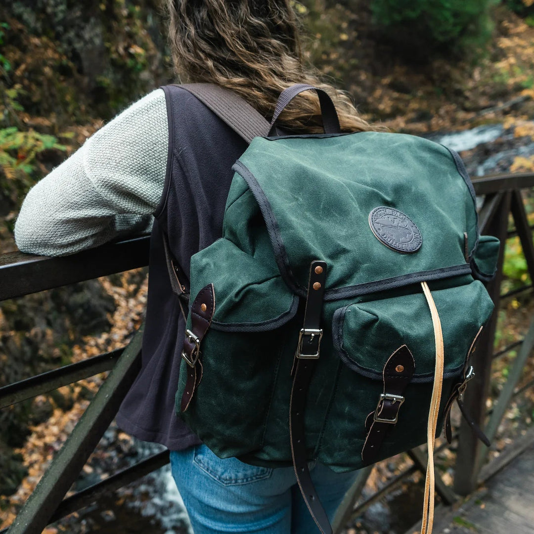 A woman on a rustic bridge in a brown vest wearing a waxed olive drab Gitchi Gammi Duluth Pack wanderer hiking backpack with a river and autumn leaves in the background.